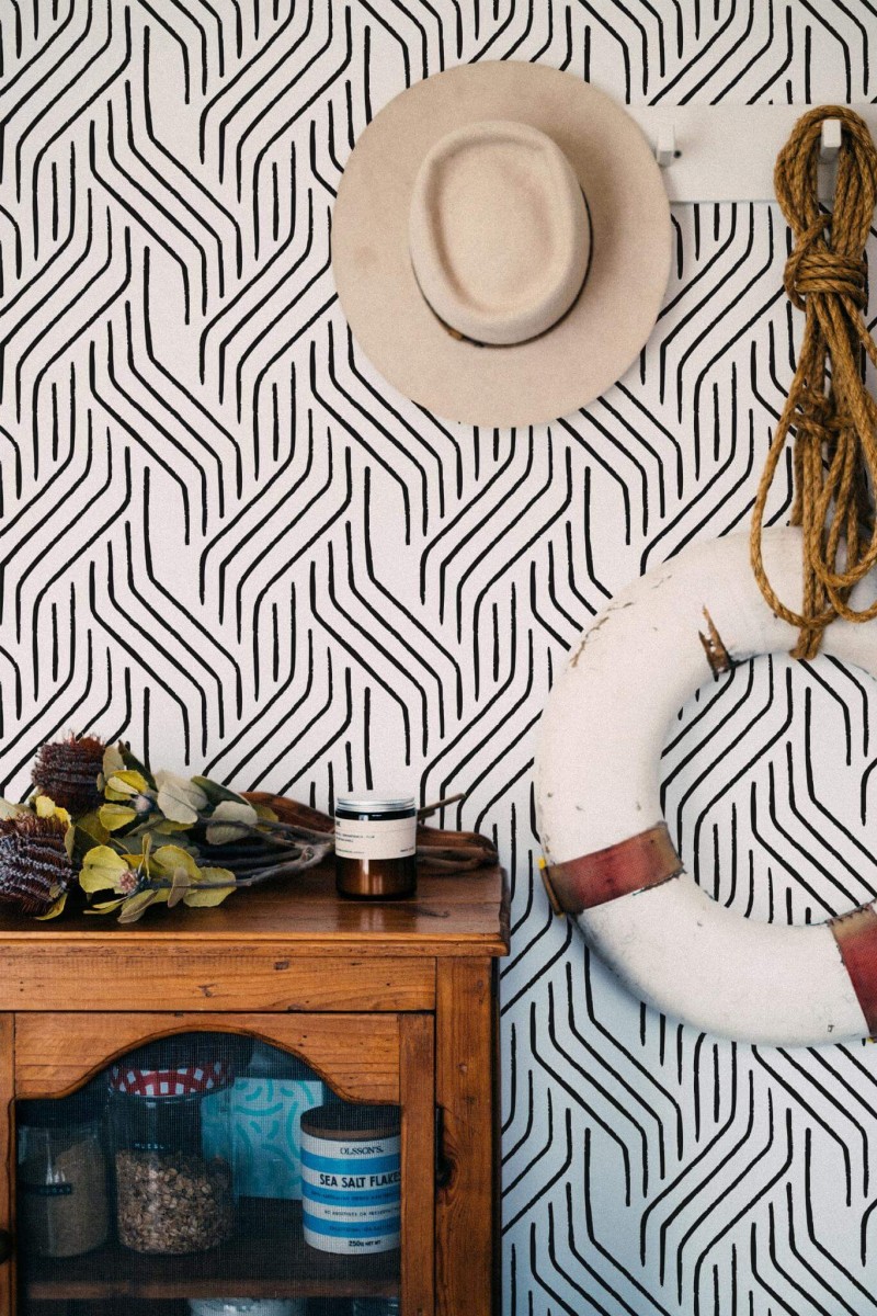 Seamless Modern Brushstroke wallpaper with bold black brush lines forming a dynamic, abstract pattern on a white background, styled in a coastal-inspired nook featuring a rustic wooden cabinet, a hanging lifebuoy, rope accents, and a neutral-toned hat. The striking black-and-white design adds a modern, artistic flair to the space.