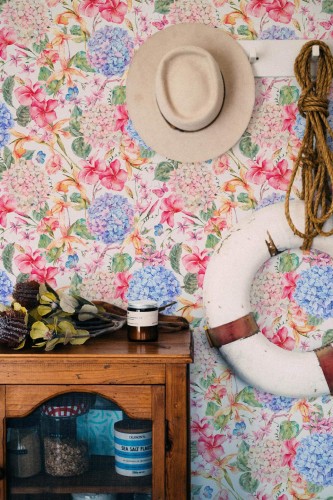 Bright Floral Wallpaper featuring watercolor-style pink, blue, and purple hydrangeas and lilies with green leaves on a white background. The botanical pattern adds a cheerful, painterly touch and vintage charm to the wall, styled with a rustic wood cabinet, coastal rope, and neutral accessories for a relaxed, nature-inspired interior.