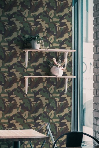 Green and brown camouflage wallpaper featuring an abstract military pattern decorates a modern interior wall. Two light wooden floating shelves with white brackets hold potted plants with cascading greenery and flowers. A minimalist light-wood table with a black metal frame and a metal chair sits below the wallpaper. The scene is softly lit by natural light coming through a tall window with teal framing, giving the space a relaxed and nature-inspired ambiance. Ideal for cafés, creative studios, or bold hallway décor, this wallpaper is shown in a matte finish and customizable size.