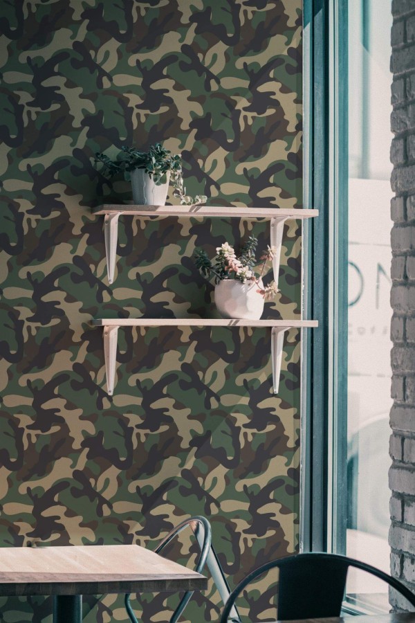 Green and brown camouflage wallpaper featuring an abstract military pattern decorates a modern interior wall. Two light wooden floating shelves with white brackets hold potted plants with cascading greenery and flowers. A minimalist light-wood table with a black metal frame and a metal chair sits below the wallpaper. The scene is softly lit by natural light coming through a tall window with teal framing, giving the space a relaxed and nature-inspired ambiance. Ideal for cafés, creative studios, or bold hallway décor, this wallpaper is shown in a matte finish and customizable size.