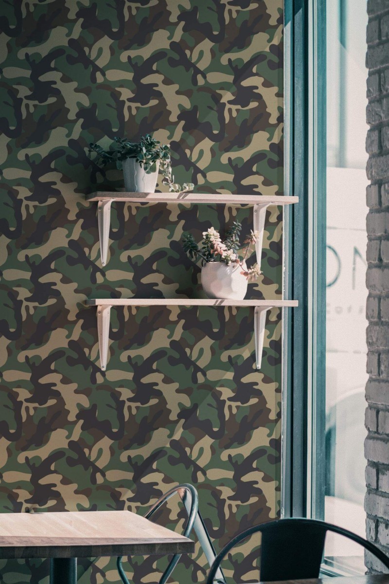 Green and brown camouflage wallpaper featuring an abstract military pattern decorates a modern interior wall. Two light wooden floating shelves with white brackets hold potted plants with cascading greenery and flowers. A minimalist light-wood table with a black metal frame and a metal chair sits below the wallpaper. The scene is softly lit by natural light coming through a tall window with teal framing, giving the space a relaxed and nature-inspired ambiance. Ideal for cafés, creative studios, or bold hallway décor, this wallpaper is shown in a matte finish and customizable size.