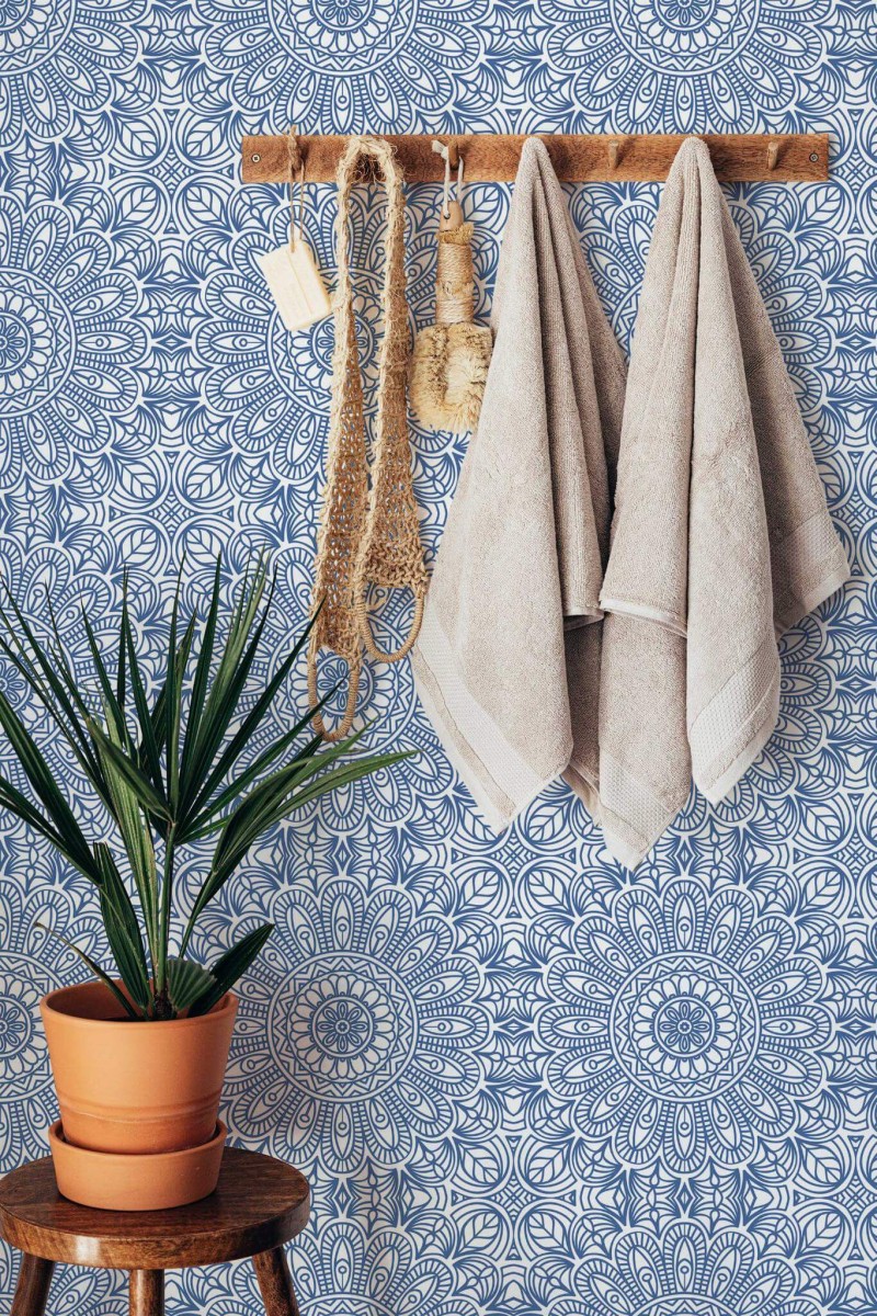 Blue mandala wallpaper serving as a striking backdrop with intricate, repeating circular patterns. A rustic wooden rack mounted on the wall holds rolled jute rope, beige towels, and a crocheted macramé hanger, adding natural textures. Below, a small wooden stool supports a terracotta planter containing a leafy green houseplant. The combination of bohemian décor elements and detailed mandala motifs creates a serene, stylish corner perfect for a bathroom or entryway.