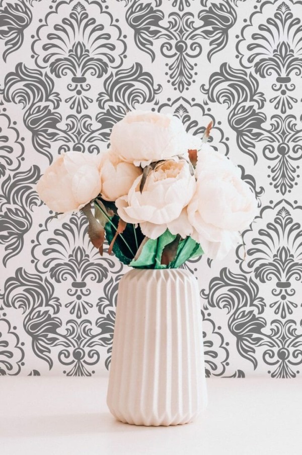 Damask patterned vintage wallpaper in soft gray and white tones adds refined elegance to a mid-century style living room. The wallpaper's ornamental floral motifs echo historic French interiors, while modern furnishings like a tan leather sofa, black-and-white woven cushion, and sculptural brass reading lamp create contrast. A tall green fiddle leaf fig in a white planter brings a touch of nature to the calm, curated interior.
