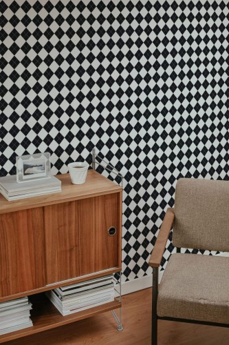A bold black and white Harlequin wallpaper featuring a high-contrast diamond grid pattern with subtle white dotted accents inside each black diamond. The design adds rhythm and structure to the space, pairing elegantly with mid-century modern furniture including a walnut sideboard, neutral armchair, and stacked books. The Fancy Walls peel and stick or non-pasted wallpaper offers a sophisticated visual texture ideal for bathrooms, home offices, or statement accent walls.