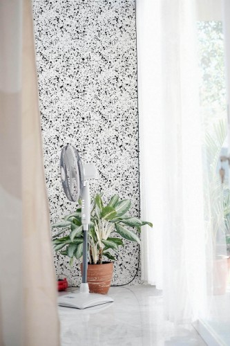 Modern sunlit interior featuring a minimalist black and white paint splatter wallpaper from Fancy Walls, used on a small accent wall next to a floor-to-ceiling window with sheer curtains. The bold abstract pattern adds visual texture to the bright, airy space. A standing fan and leafy potted plant bring natural balance to the clean lines and polished marble floor. This wallpaper is available in peel and stick or non-pasted formats, printed with eco-safe GreenGuard Gold certified inks on PVC-free materials.