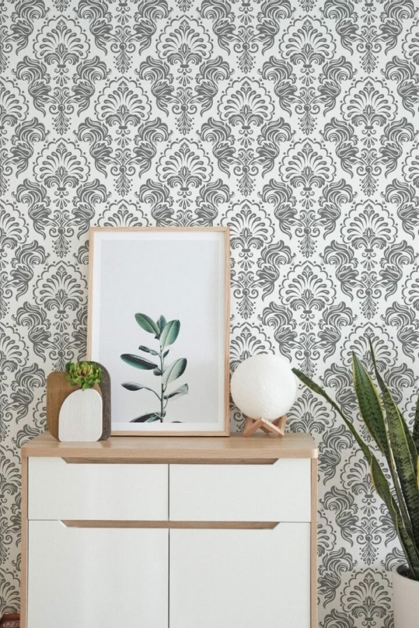 Mid-century modern living room styled with gray damask peel and stick wallpaper featuring an elegant symmetrical floral pattern on white background. The ornate vintage wallpaper design creates a timeless Art Deco look, paired with a caramel leather armchair, a sculptural gold lamp, and a large potted fiddle leaf fig. The setup includes a white radiator and neutral textiles, blending cozy warmth with classic sophistication.