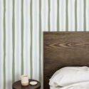 Wavy Sage Stripes peel and stick wallpaper with soft sage and light blue vertical lines behind a dark wood headboard.