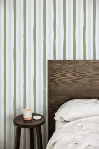 Wavy Sage Stripes peel and stick wallpaper with soft sage and light blue vertical lines behind a dark wood headboard.