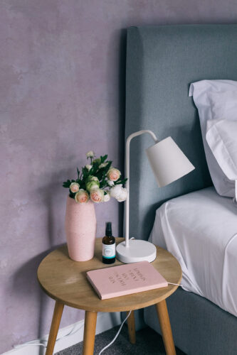 Cloudy Pink Plaster Look peel and stick wallpaper as a soft textured backdrop in a bedroom beside a bed and lamp.