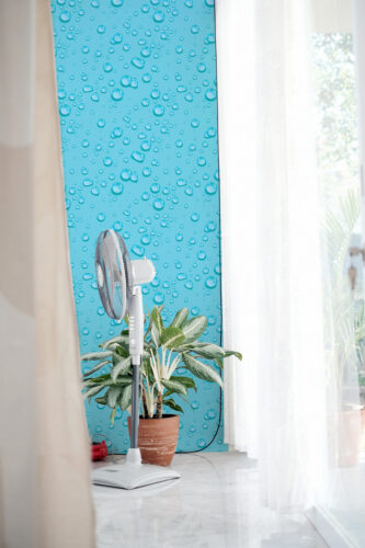 Dense Turquoise Raindrops peel and stick wallpaper with glossy water droplet pattern on a turquoise wall beside sheer curtains.