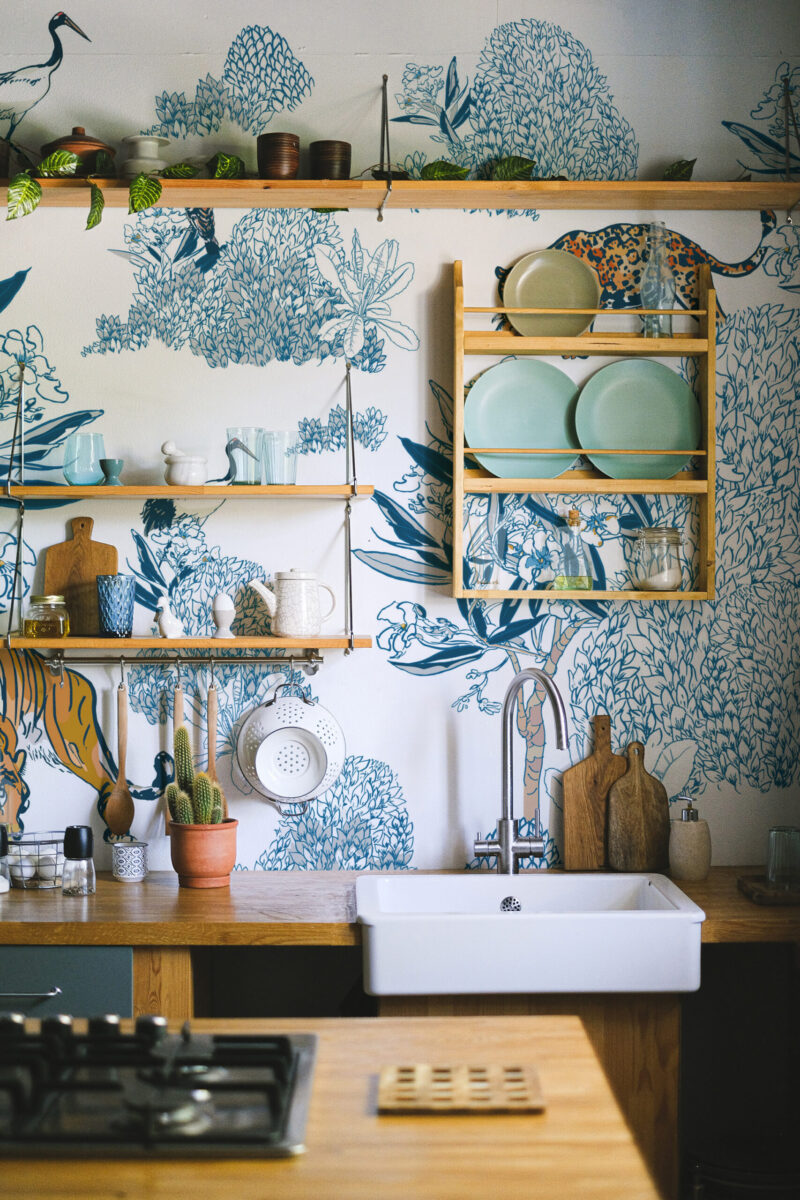 Chinoiserie Big Cats and Birds peel and stick wallpaper mural in a kitchen, with blue botanical drawings, cranes, and spotted big cats behind open shelves.