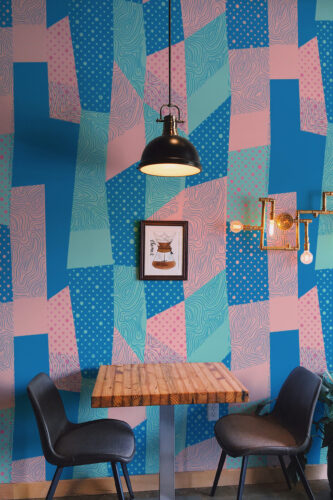 Retro Geometric Patchwork shows a full-wall wall mural of blue and pink abstract patchwork shapes behind a small table.