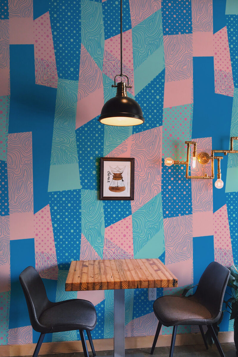 Retro Geometric Patchwork shows a full-wall wall mural of blue and pink abstract patchwork shapes behind a small table.