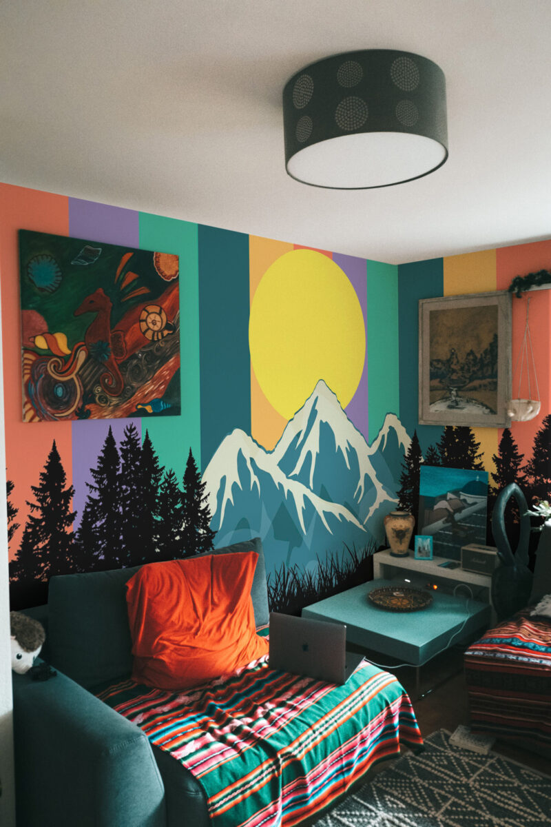 Rainbow Mountain Sunrise wall mural wrapping a bedroom corner with striped sky, yellow sun, snowy peaks, and pine silhouettes.
