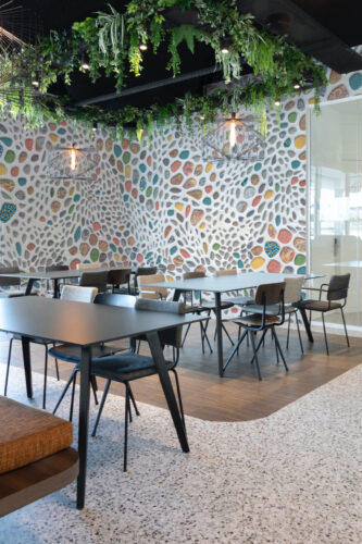 Colorful Pebble Mosaic Look wall mural covers a full wall in a modern café seating area with hanging greenery overhead.