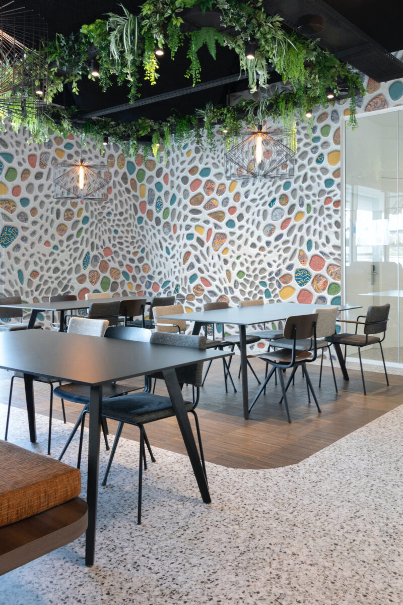 Colorful Pebble Mosaic Look wall mural covers a full wall in a modern café seating area with hanging greenery overhead.