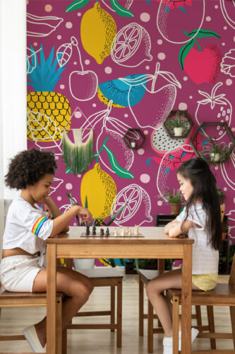 Tropical Fruit Doodles peel and stick wallpaper mural with bold fruit sketches on a magenta background behind a kids table and chairs.