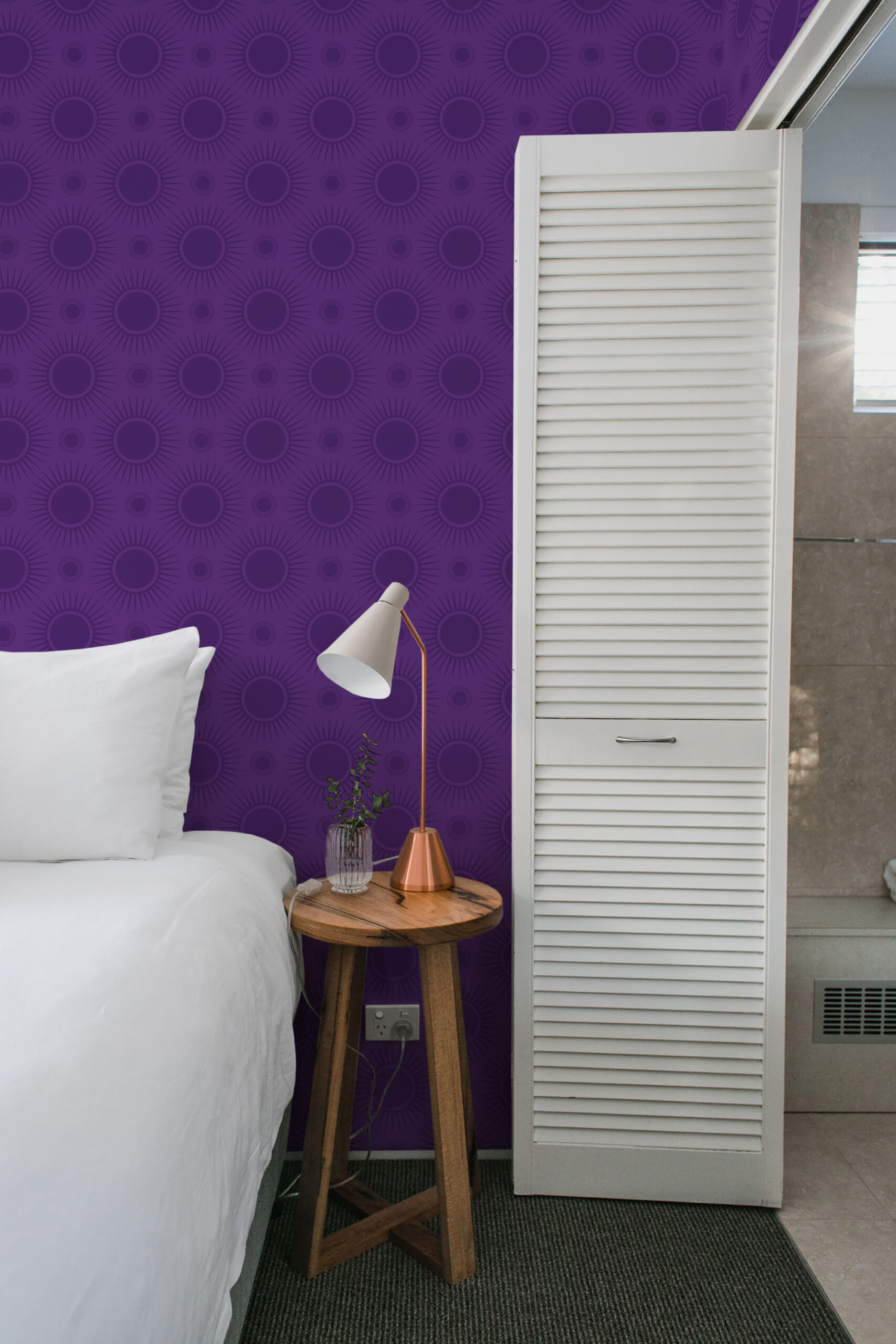 Bold Purple Sunburst pre-pasted wallpaper creates an accent wall of repeating sunburst circles behind a bedside table.