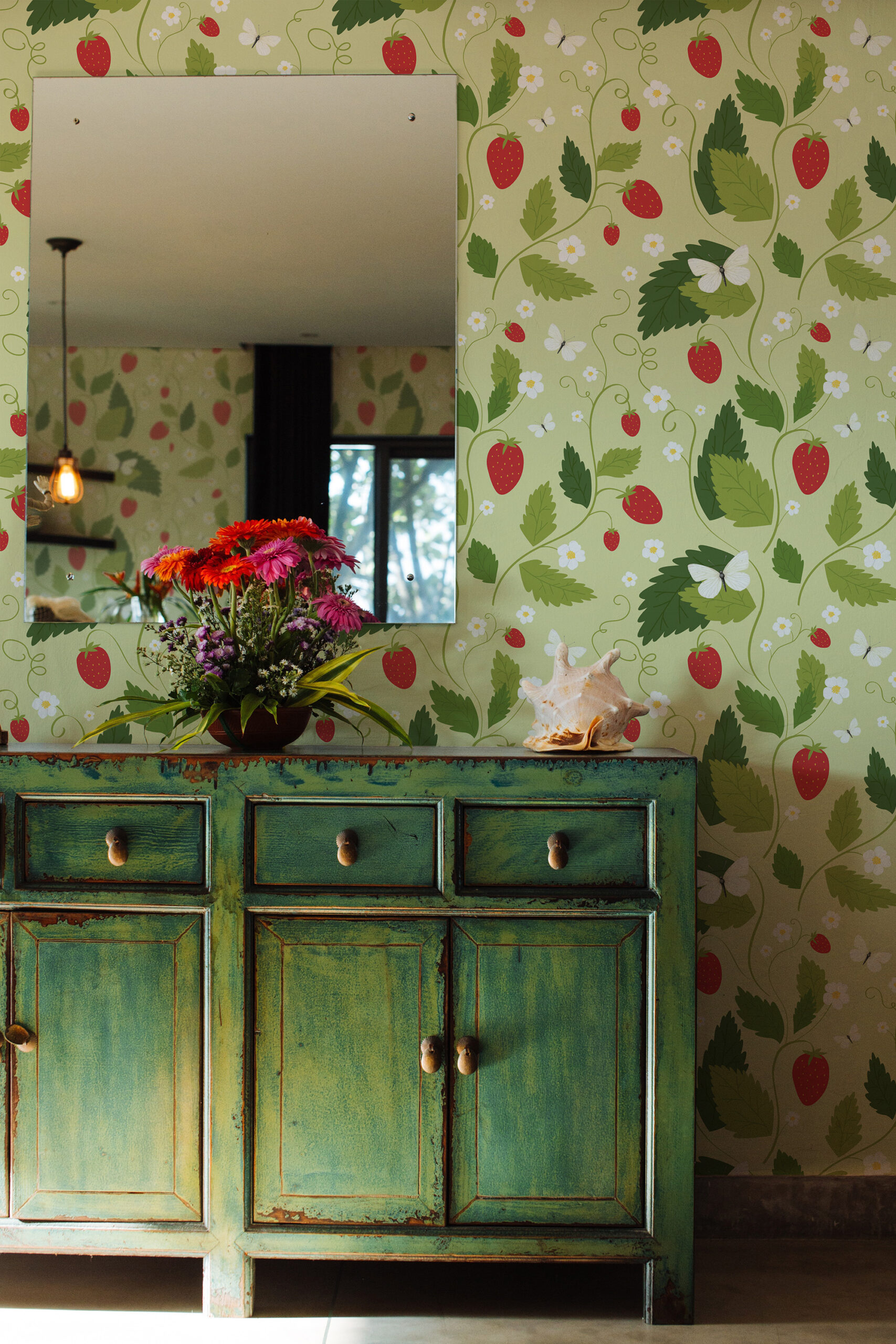 Cute, whimsical, strawberry stick wallpaper with red strawberries, green leaves, and small white flowers behind a wall mirror.