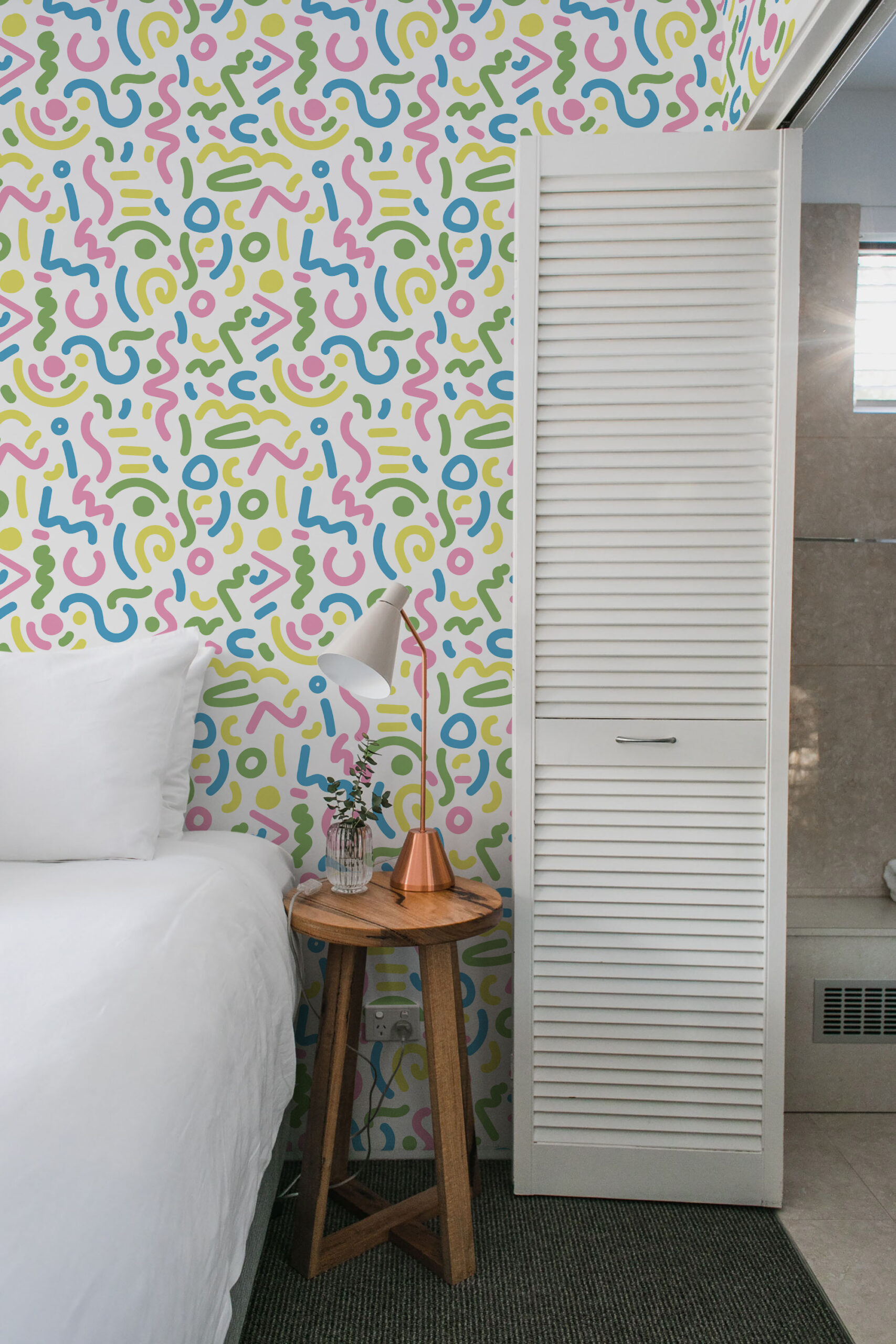 Bold White Squiggle pre-pasted wallpaper with pink, blue, and green doodle lines on an accent wall by the bed.