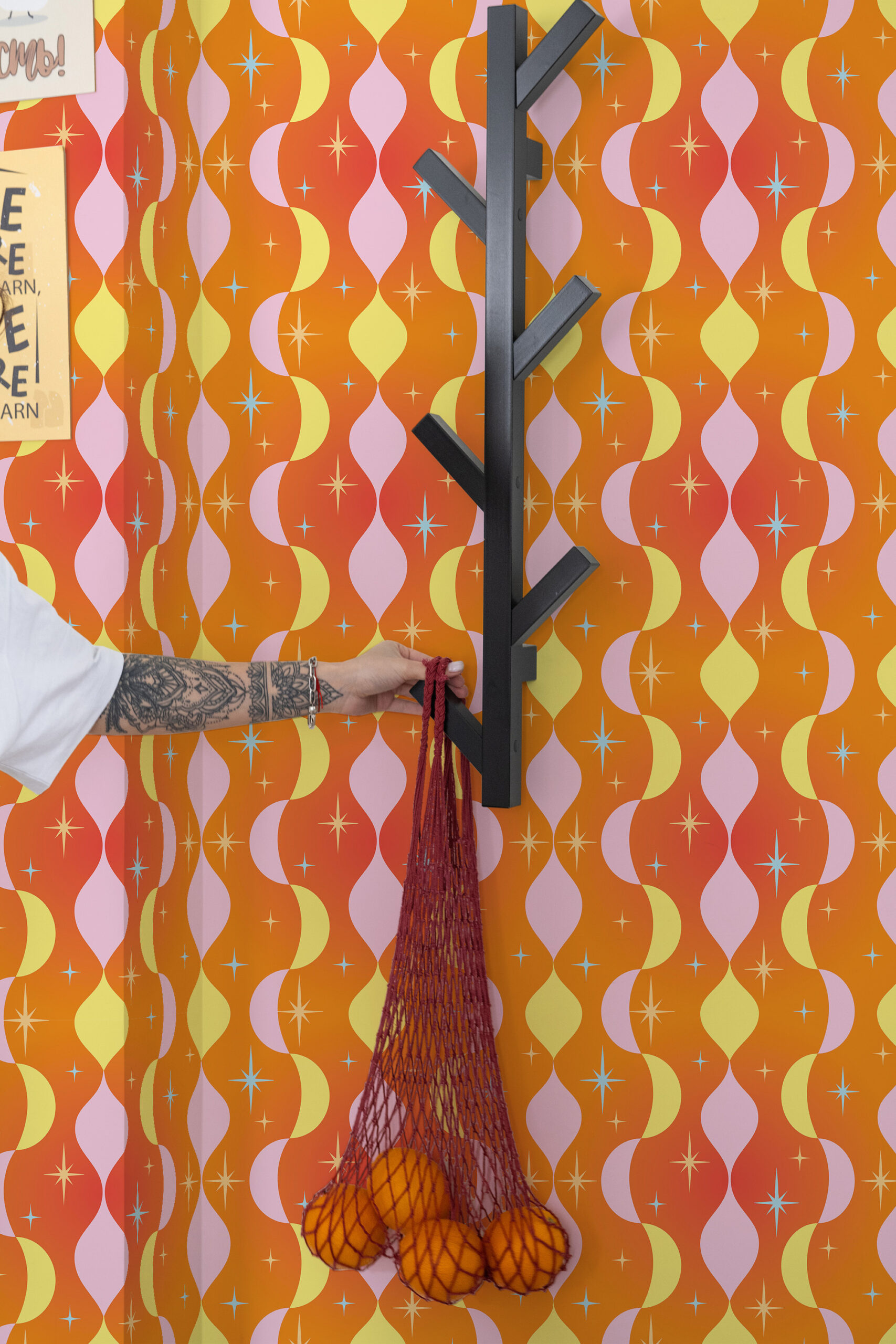 Retro Orange Stars peel and stick wallpaper with wavy orange, yellow, and pink stripes and small starbursts behind a wall hook.