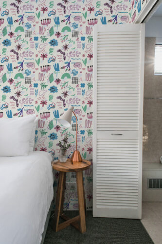 Playful Ivory Abstract Shapes peel and stick wallpaper with colorful doodle-style shapes on a narrow bedroom wall beside the bed.