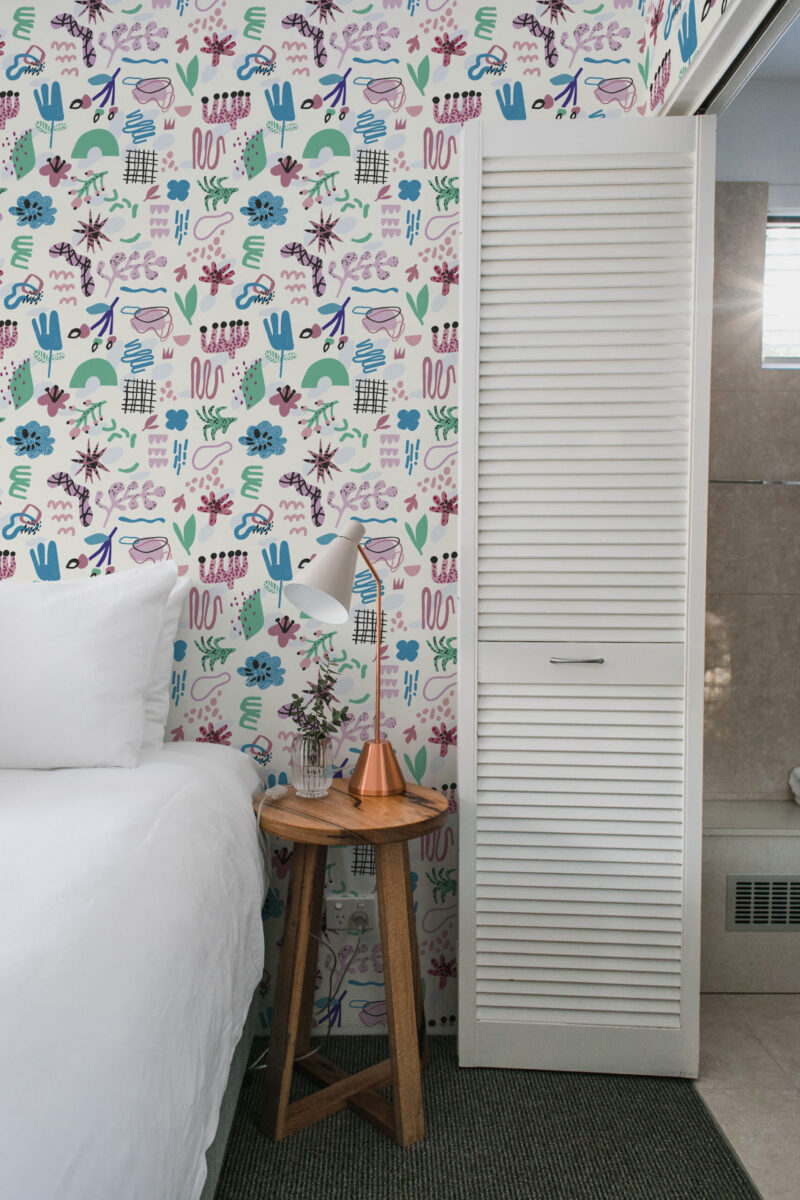 Playful Ivory Abstract Shapes peel and stick wallpaper with colorful doodle-style shapes on a narrow bedroom wall beside the bed.