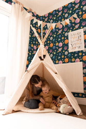 Dense Teal Space peel and stick wallpaper with colorful planets, rockets, and stars covering a kids’ playroom wall.