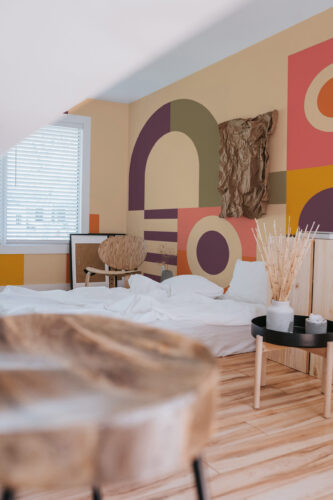 Retro Geometric Playroom peel and stick wall mural with bold arches and circles in muted pink, olive, purple, and orange behind a bed.