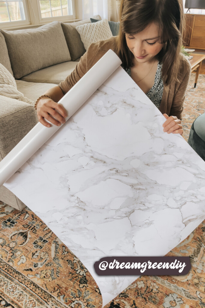 Person examining a roll of white marble-patterned peel-and-stick wallpaper in a cozy living room