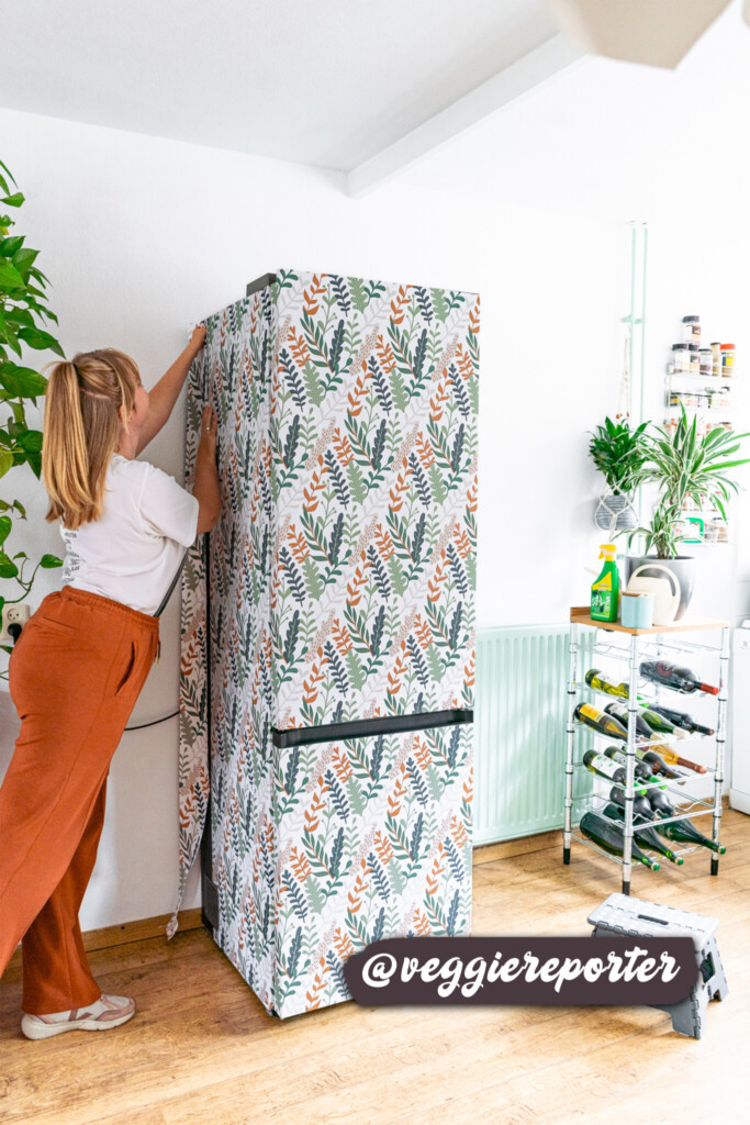 A refrigerator wrapped in a vibrant botanical wallpaper with orange, green, and gray leaves, adding a fresh and stylish touch to a modern kitchen surrounded by lush indoor plants