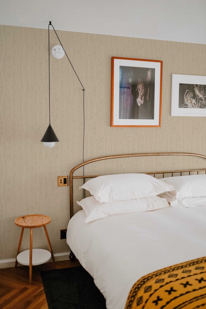 Warm bedroom with a black pendant light, framed artwork, and curved metal bed frame, set against Sand Retro Talbott Grasscloth wallpaper. The soft beige weave effect introduces texture and comfort. Fancy Walls removable peel and stick or non-pasted wallpaper.