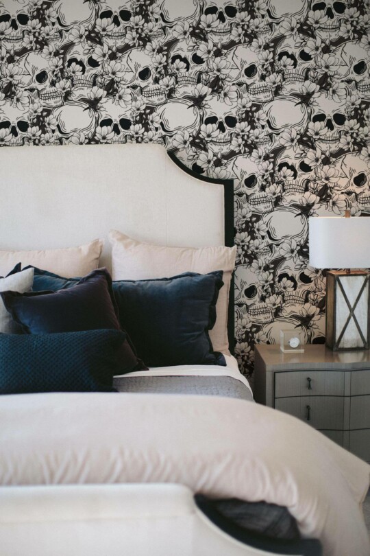 Black and White Skull Wallpaper featuring intricately detailed floral and skull motifs, styled behind a white upholstered bed with plush navy pillows and a gray nightstand holding a lantern-style lamp. The dramatic monochrome pattern adds edgy elegance to the cozy bedroom décor.