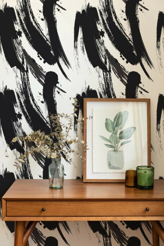 The bold black brush strokes of this wallpaper create a striking, dynamic statement wall in any room. The sweeping strokes, varying in thickness and intensity, evoke an artistic, expressive feel, blending abstract design with a sense of fluid motion. The high contrast of black on a neutral background adds visual depth, making it a perfect fit for modern interiors. This wall features a minimalist, Scandinavian-inspired design with a natural wood table, framed art, and subtle green plants, complementing the wallpaper’s bold artistic theme. The Fancy Walls peel and stick or non-pasted wallpaper brings effortless style and eco-conscious design to your space.