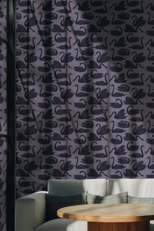 Modern accent wall featuring black swan wallpaper with hand-drawn swans in deep black and subtle navy, arranged on a muted purple background. The graceful swan pattern adds elegance and movement, ideal for artistic interiors. A cozy corner sofa with textured gray cushions and a round wooden coffee table creates a warm, inviting seating area. Natural light and soft shadows enhance the bold, moody atmosphere of the room.