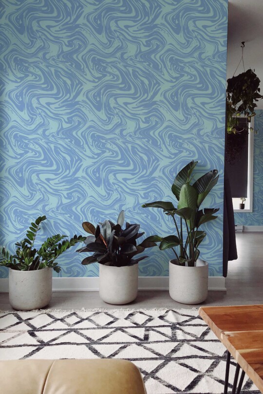 A modern living area featuring a swirling blue marble-patterned wallpaper as the accent wall. The wallpaper design shows flowing, water-like swirls in varying shades of sky, aqua, and navy blues, adding a serene, abstract aesthetic. Three large concrete planters filled with lush tropical houseplants sit in front of the wall, complemented by a geometric rug and simple wooden coffee table. The overall style is contemporary and airy, with clean lines and subtle decorative elements that highlight the calming, fluid wallpaper design.