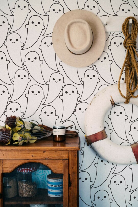 Cute black and white ghost pattern wallpaper decorates an eclectic hallway with vintage nautical accents and a rustic wooden cabinet. The charming, friendly ghost illustrations on a crisp white background create a whimsical and playful mood, ideal for accent walls in kids' rooms or quirky interiors. Styled with a beige felt hat, rope, and retro glass jars, this design adds a cheerful twist to natural wood and neutral tones. Made with Fancy Walls peel and stick or non-pasted wallpaper for easy application and removal.