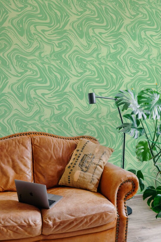 A living room scene with a swirling green marble-patterned wallpaper behind a vintage-style brown leather sofa. A laptop rests on the sofa, and a tall black floor lamp stands to the side. A large leafy plant adds a fresh, nature-inspired accent to the modern interior.