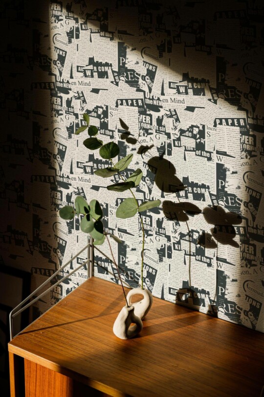 Black and white newspaper wallpaper featuring vintage-style headlines and fragmented text clippings, adding a bold, eclectic look to a modern interior corner. The wall is partially bathed in warm natural light, casting leafy plant shadows across the design. A wooden cabinet topped with a sculptural ceramic vase and eucalyptus stems sits in front, enhancing the room's artistic and organic vibe. Made by Fancy Walls, the wallpaper is available in peel and stick or non-pasted PVC-free formats, printed with GreenGuard Gold certified inks.