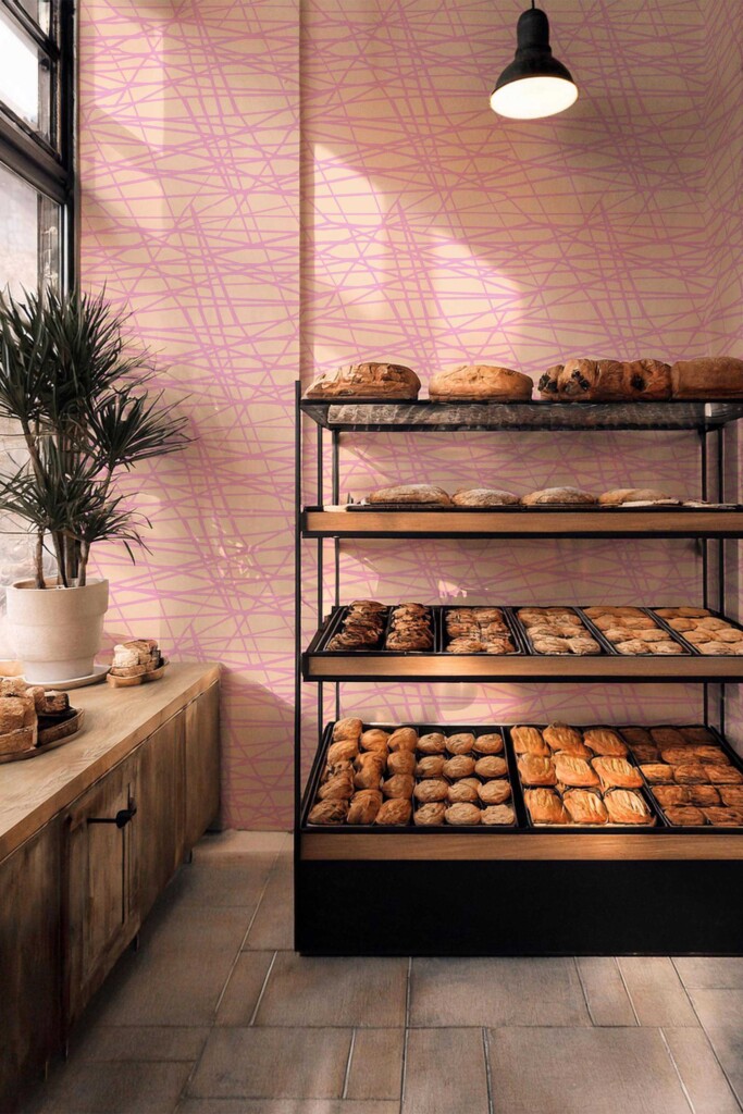A bakery interior with sunlight warming the space, featuring Pink Bakery Linear Vibes wallpaper by Fancy Walls peel and stick or non-pasted wallpaper. The soft peach background with overlapping pink linear strokes brings movement and charm to cozy, eclectic spaces.