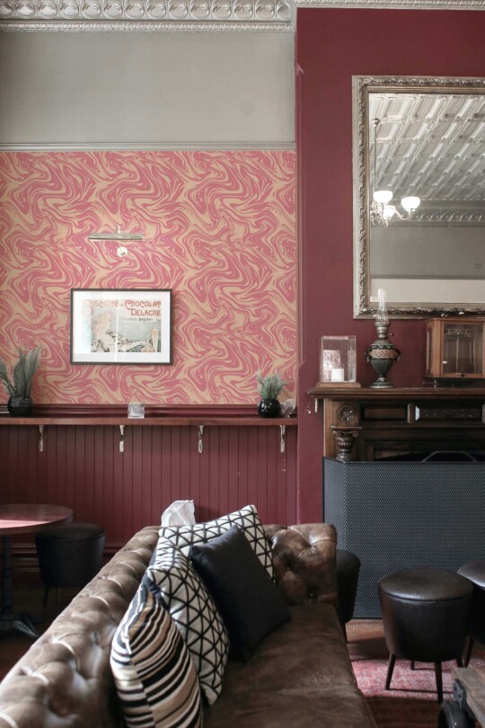 An elegant sitting room featuring Fancy Walls peel and stick or non-pasted wallpaper in a pink and cream marble swirl pattern. The wallpaper showcases flowing, abstract curves in warm blush tones layered over a soft beige background, creating a dynamic yet refined visual texture. This vintage-inspired space includes a tufted leather sofa with patterned cushions, a carved wooden mantel, and ornate ceiling moldings, all complemented by deep burgundy wall accents and antique décor. The stylish mix of old-world charm and modern artistic wallpaper results in a cozy yet sophisticated lounge atmosphere. The wallpaper is PVC-free, available in self-adhesive polyester textile or FSC mix-certified non-woven, and printed with GreenGuard Gold certified inks for vivid, safe, and eco-conscious indoor décor.