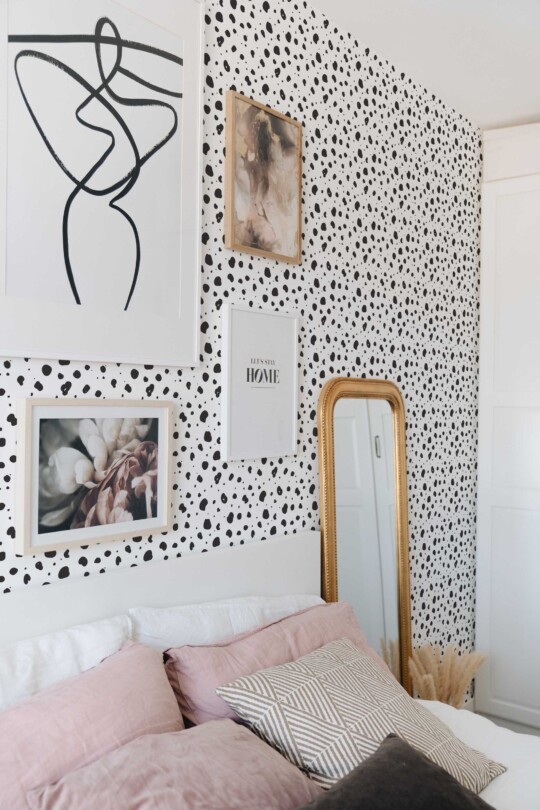 Bedroom accent wall decorated with tiny monochrome cheetah print wallpaper in black and white. A collection of framed art, including abstract line drawings and floral photography, hangs on the wall above a white headboard. Soft pink and neutral-toned pillows layer across the bed, creating a calm, modern vibe. A tall gold-framed mirror leans against the wall, reflecting the printed pattern and adding warmth. Overall, the scene feels airy, stylish, and playful.