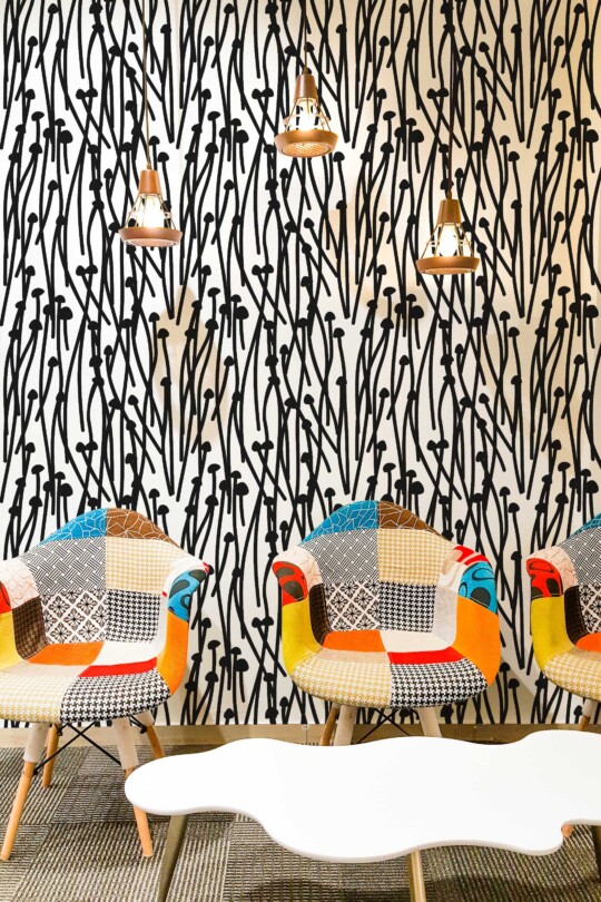 Eclectic interior featuring a bold black and white abstract wallpaper with hand-drawn tall mushrooms and wavy stems, arranged in a dense vertical pattern. The wallpaper creates a high-contrast graphic backdrop in a modern lounge space, paired with colorful patchwork armchairs in mid-century style. A white sculptural coffee table and trio of industrial copper pendant lights add warmth and balance to the vivid, artistic atmosphere. Ideal for creative spaces, cafés, or accent walls.