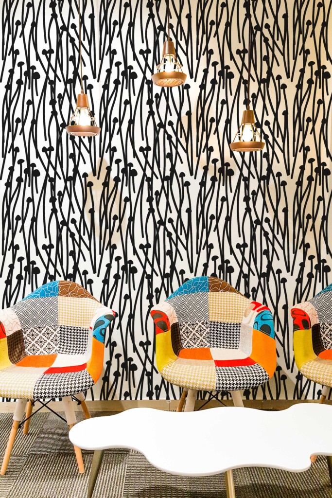 Eclectic interior featuring a bold black and white abstract wallpaper with hand-drawn tall mushrooms and wavy stems, arranged in a dense vertical pattern. The wallpaper creates a high-contrast graphic backdrop in a modern lounge space, paired with colorful patchwork armchairs in mid-century style. A white sculptural coffee table and trio of industrial copper pendant lights add warmth and balance to the vivid, artistic atmosphere. Ideal for creative spaces, cafés, or accent walls.